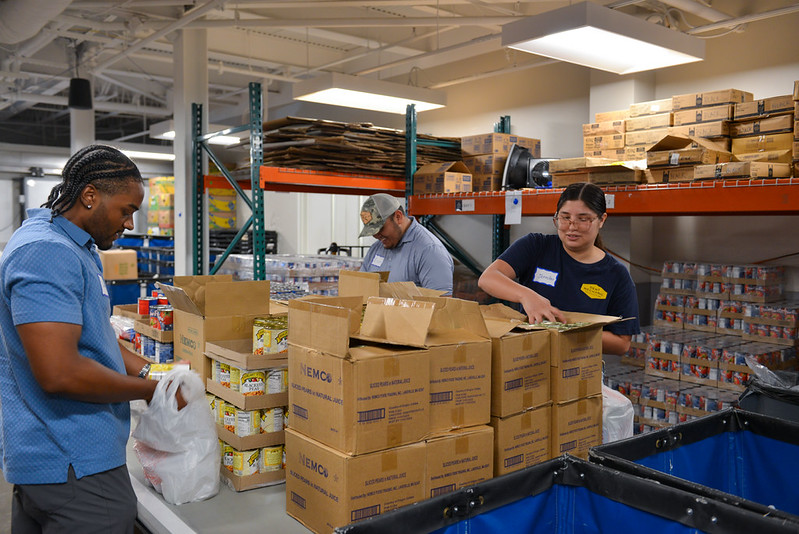 Three Dent Mechanic Group volunteers packing canned pears and other pantry items into bags inside The Storehouse warehouse.