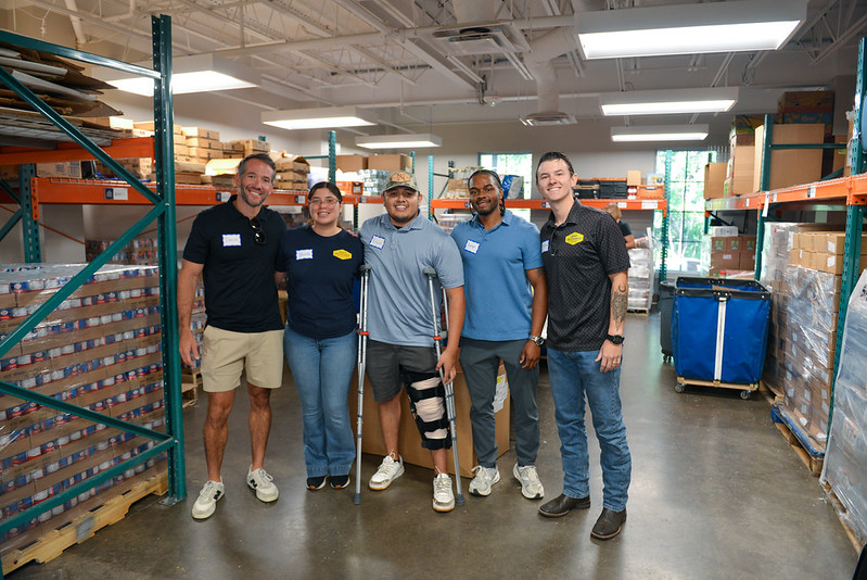 Five smiling Dent Mechanic Group teammates, some in company shirts, pose together between warehouse shelves stocked with food at The Storehouse.
